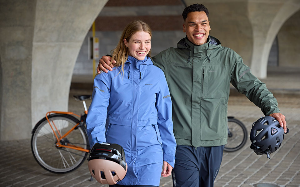 Duo souriant en veste bleue et verte avec des casques de vélo dans un passage couvert.