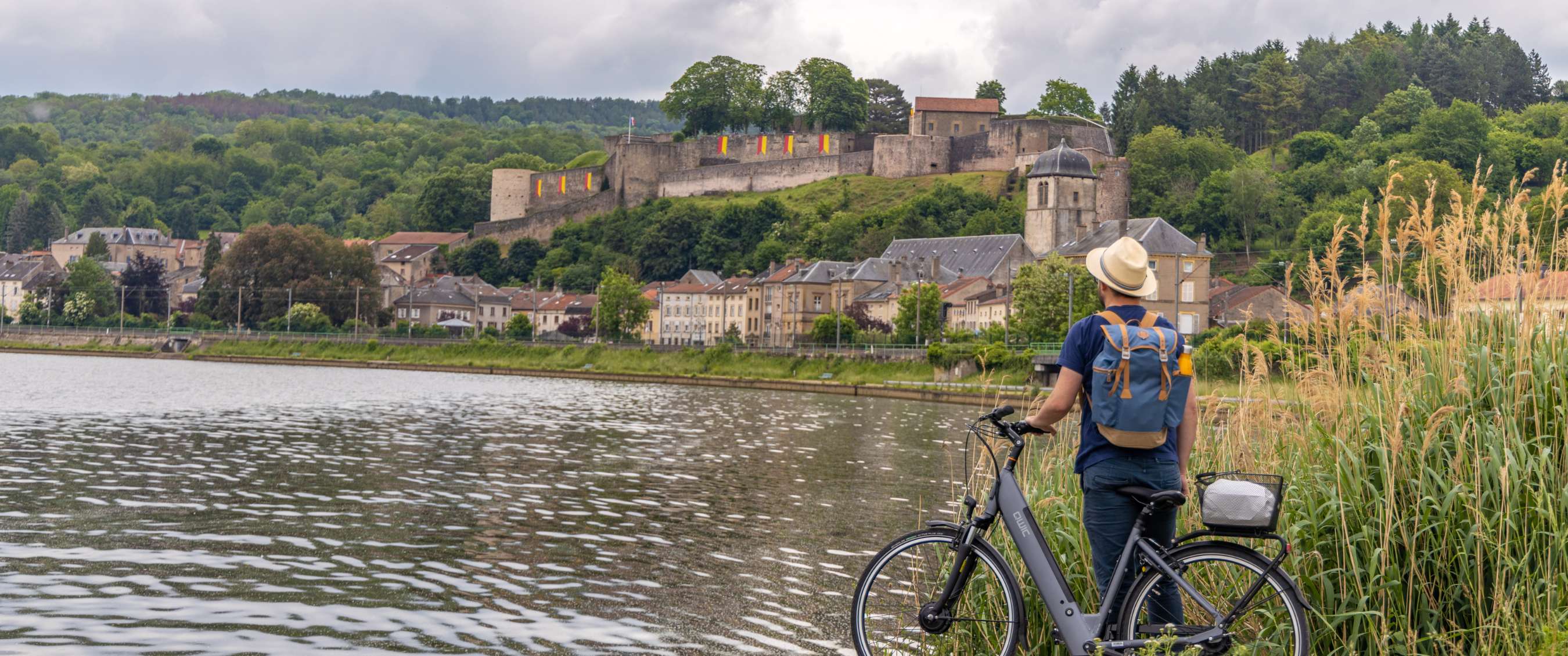 Cycliste avec vélo au bord de la rivière, face à une ville fortifiée sur une colline.