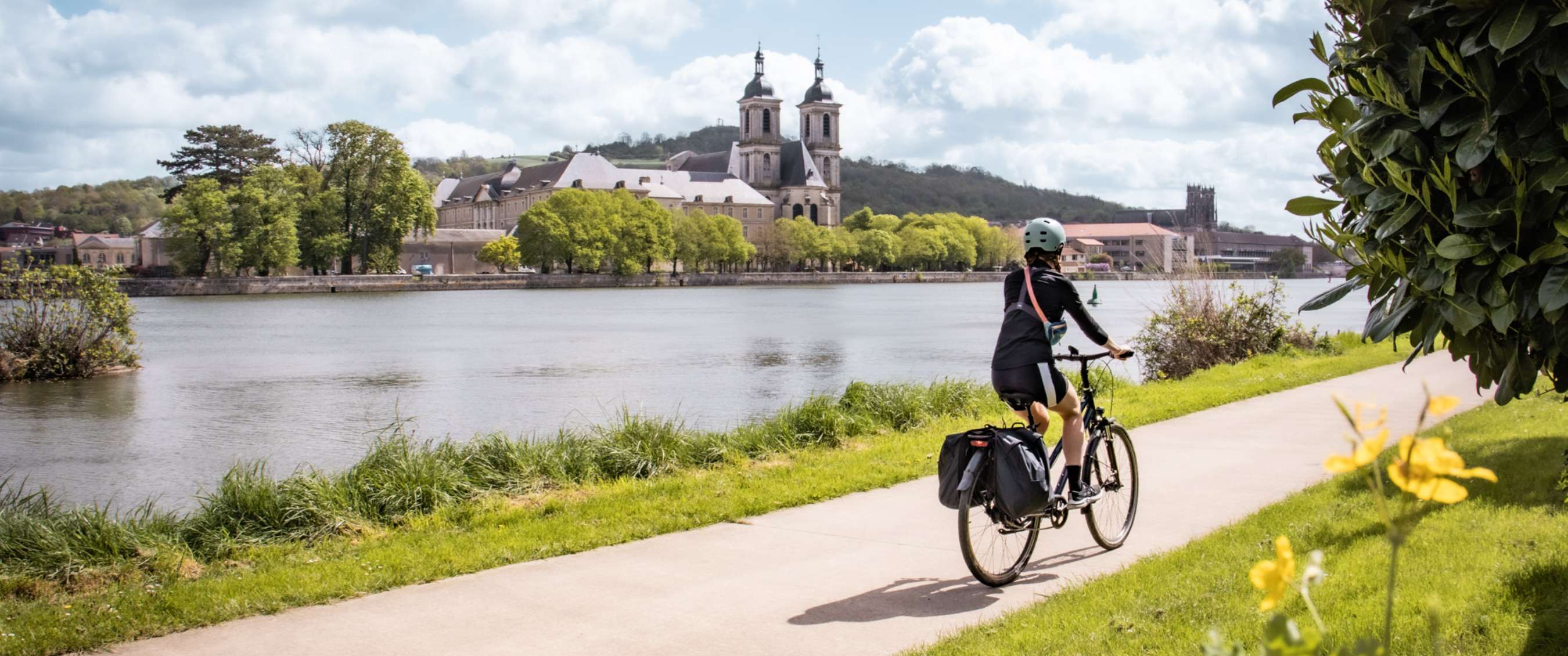 Personne à vélo sur un chemin au bord de l’eau avec une église et des arbres en face.