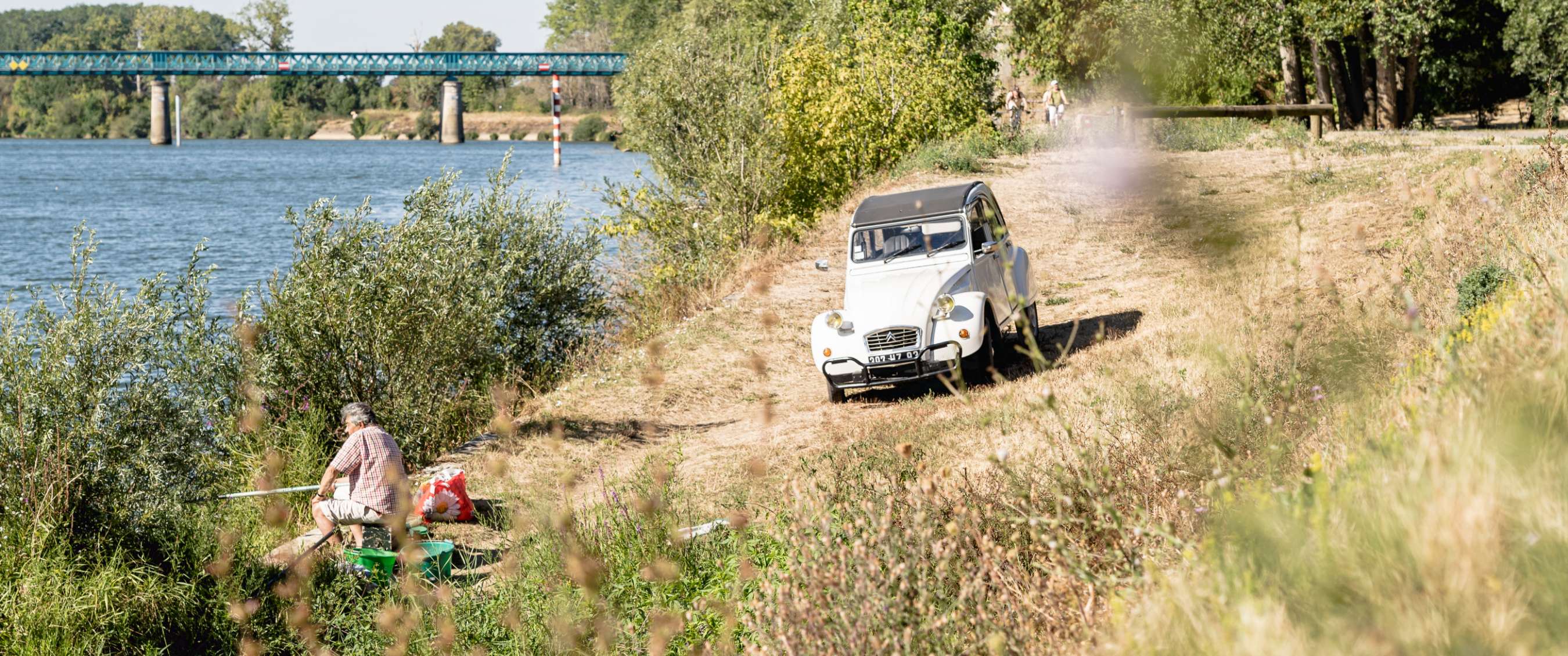 Citroën 2CV blanche sur un chemin au bord de la rivière, avec un pêcheur au premier plan.