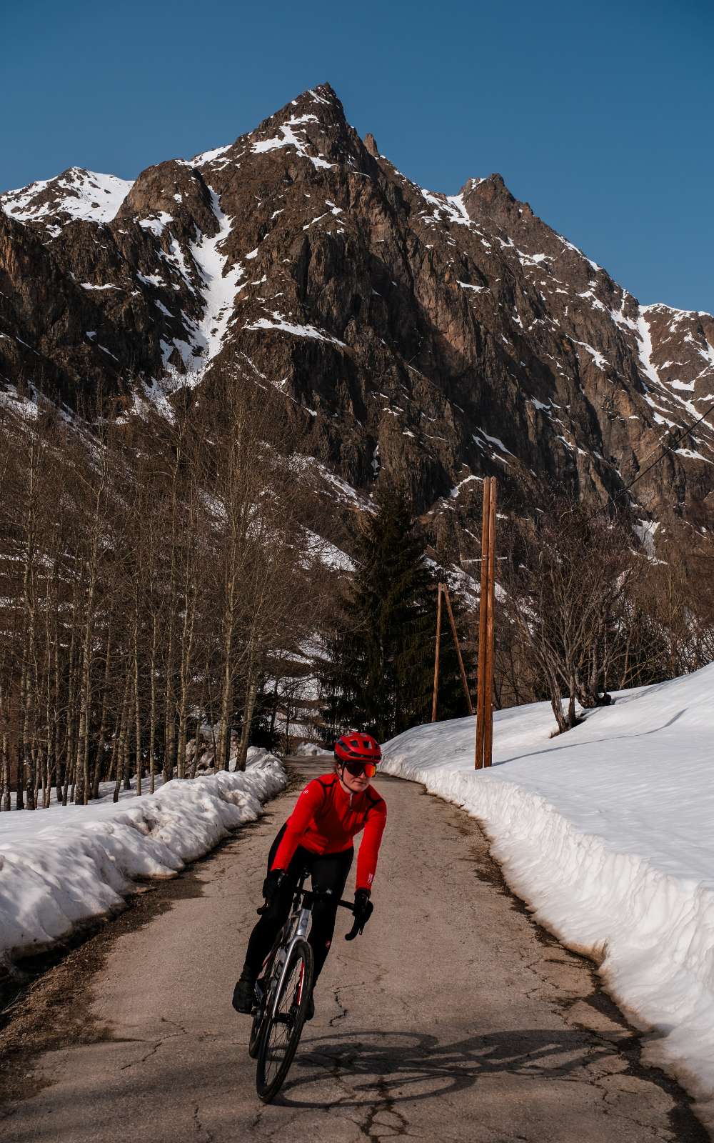 Une personne à vélo, casque et maillot rouges, roule sur une route de montagne entre des bancs de neige.