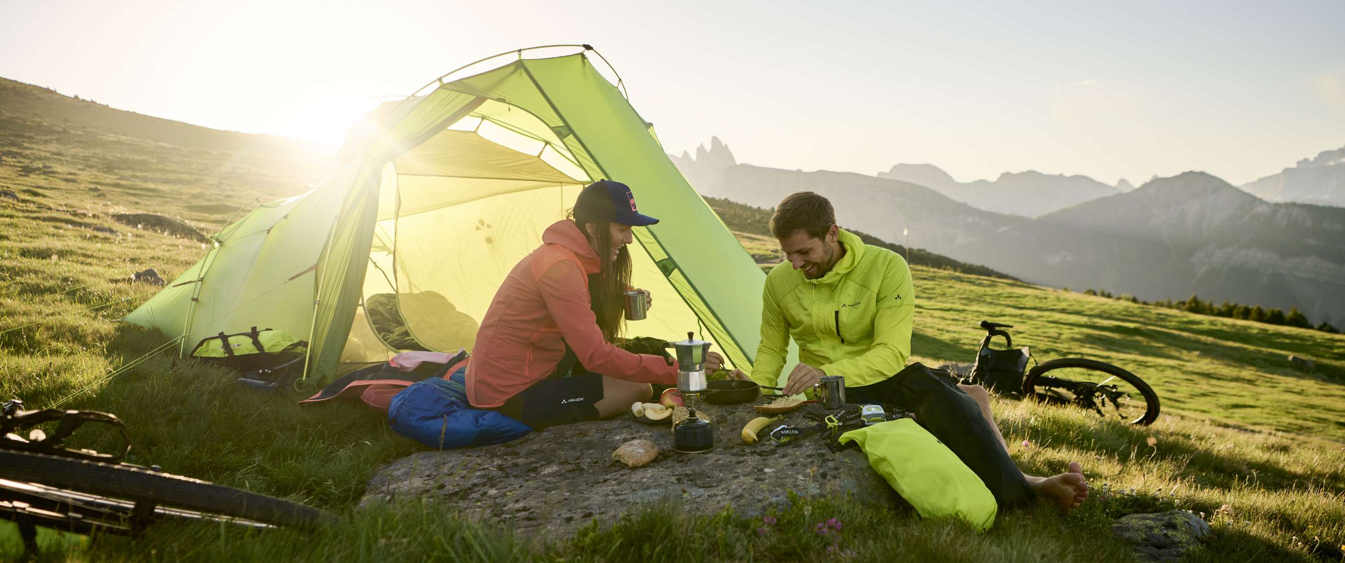 Deux personnes cuisinent près d’une tente verte sur une prairie de montagne avec des vélos à côté.