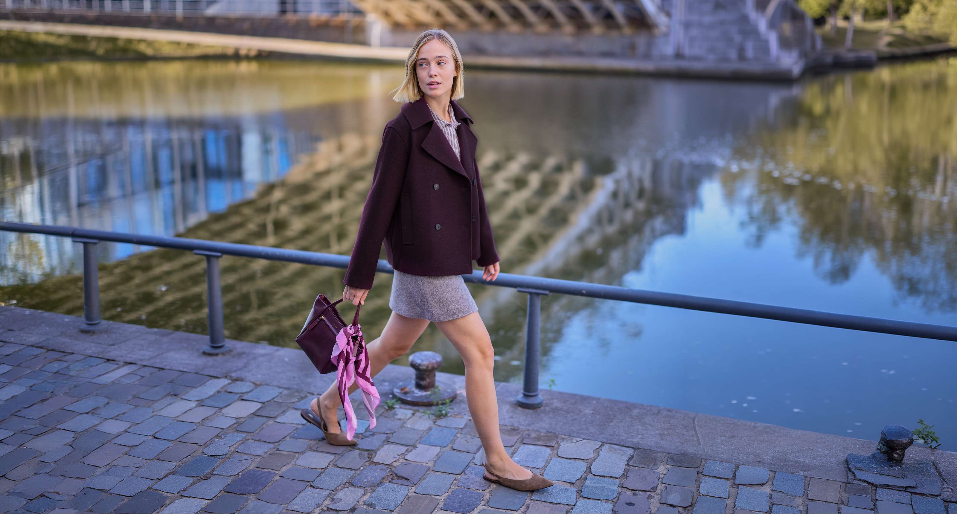 Une personne en veste bordeaux et jupe grise marche au bord de l’eau avec un sac à main.