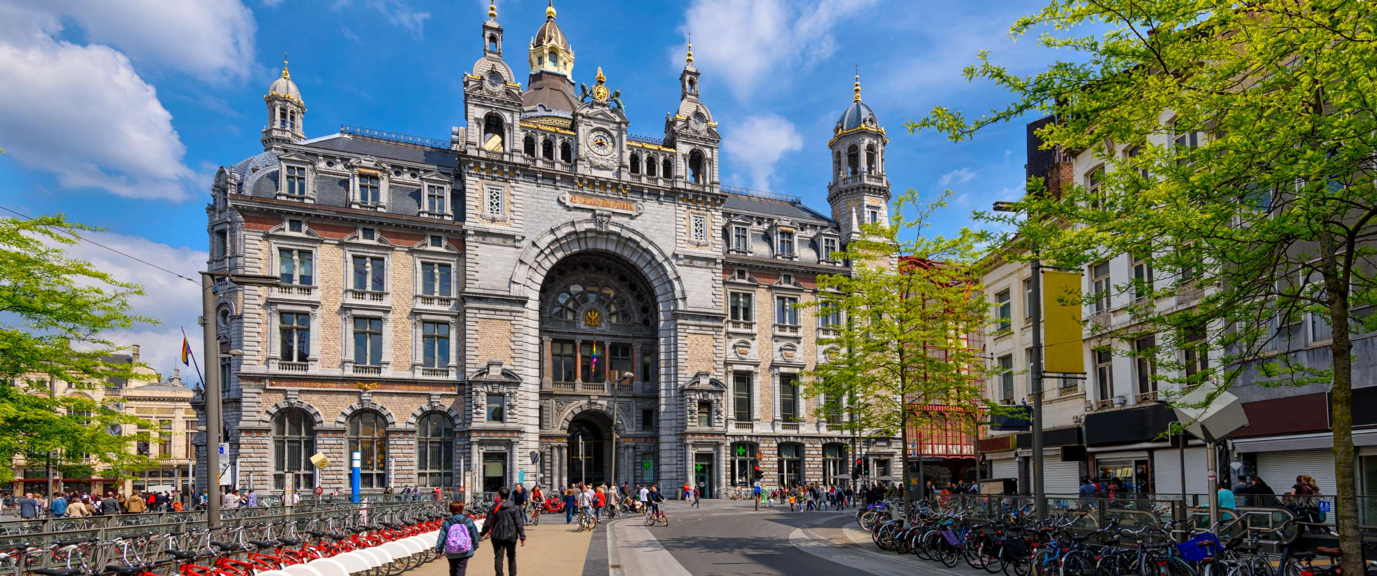 Grande façade historique de gare avec tours, vélos et passants sous un ciel bleu.