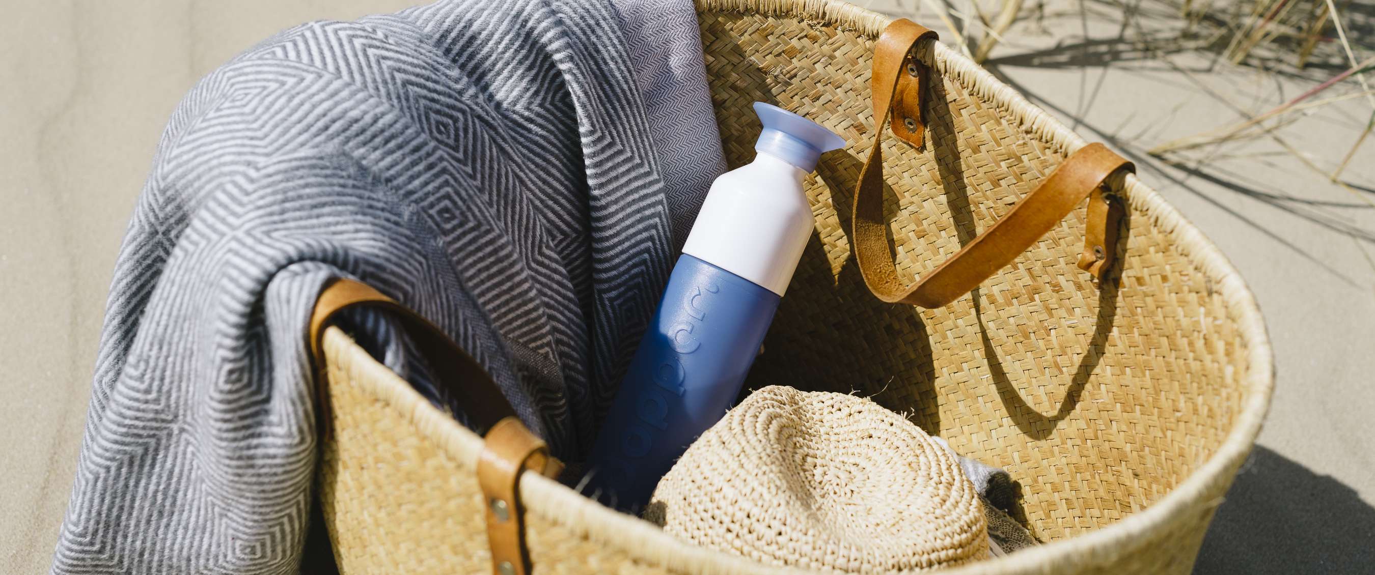 Gourde bleue Dopper dans un sac de plage tressé avec serviette et chapeau.