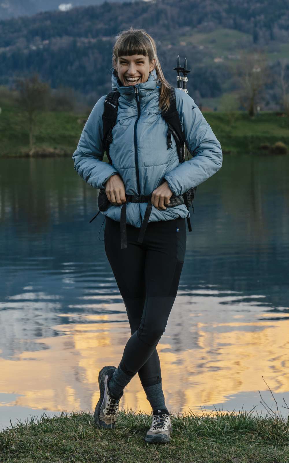 Une personne en veste matelassée et pantalon noir pose avec sac à dos devant un lac et des collines.