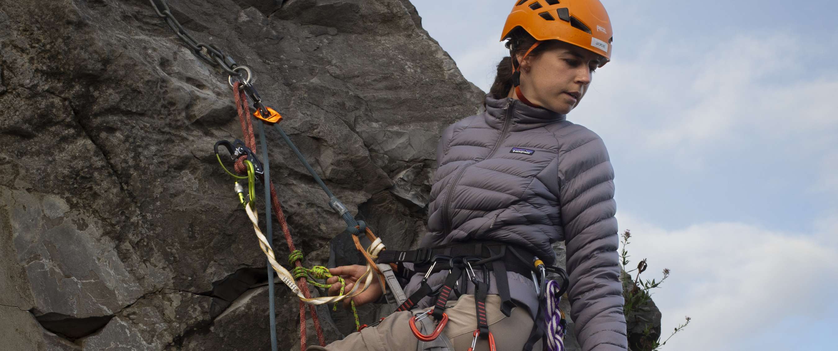 Grimpeuse avec casque orange et veste grise Patagonia assurée sur une paroi rocheuse abrupte.