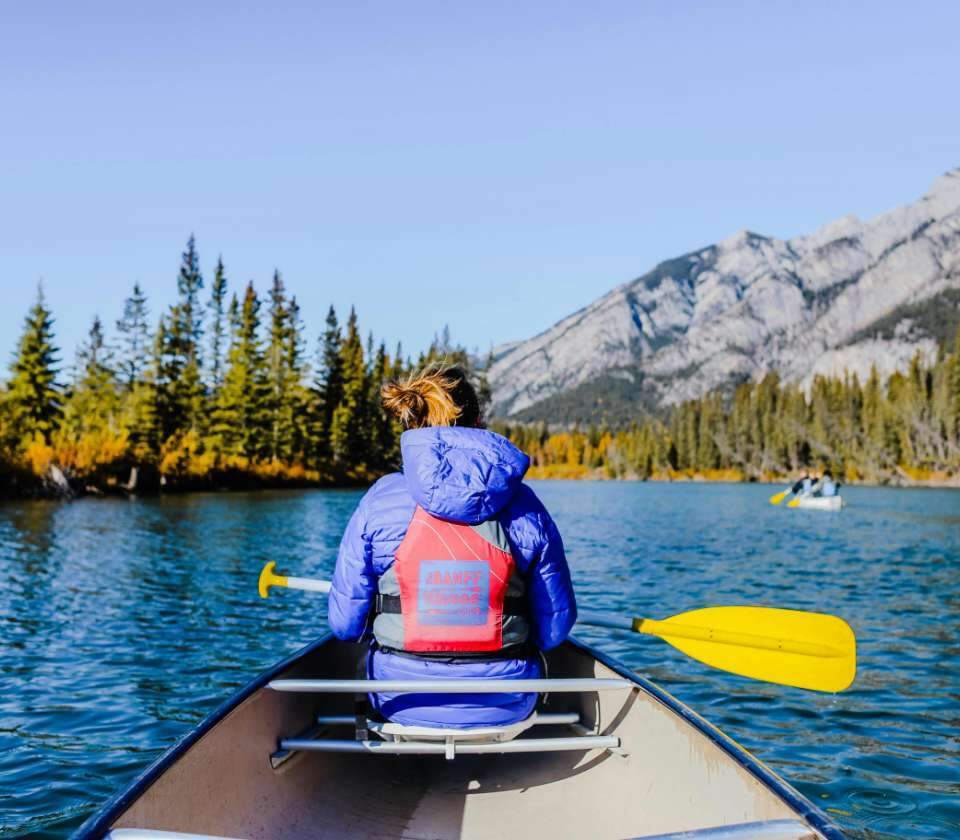 Persoon in blauwe jas vaart in een kano over een bergmeer, omringd door naaldbomen.