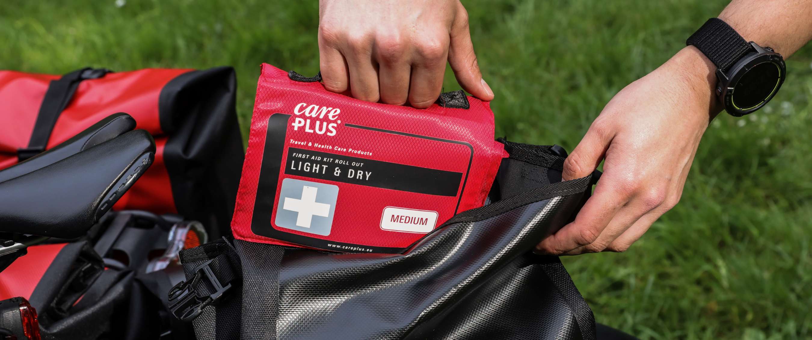 Main plaçant une trousse de secours Care Plus Light & Dry Medium dans une sacoche de vélo sur l’herbe