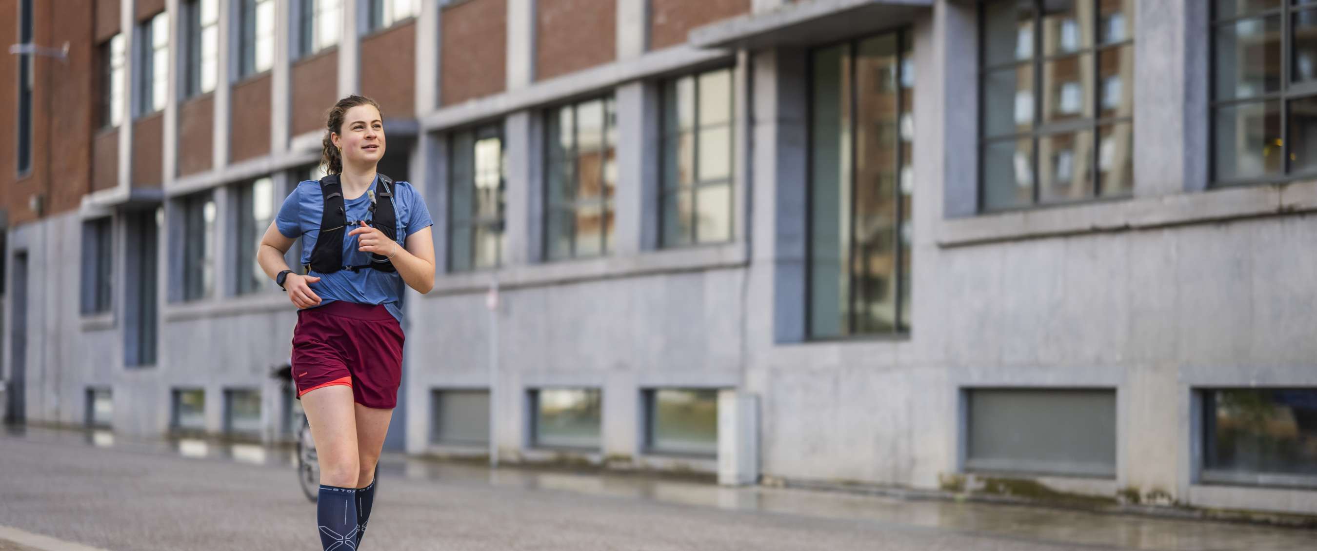 Femme courant en ville le long d’un bâtiment, avec t-shirt bleu, short bordeaux et gilet noir.