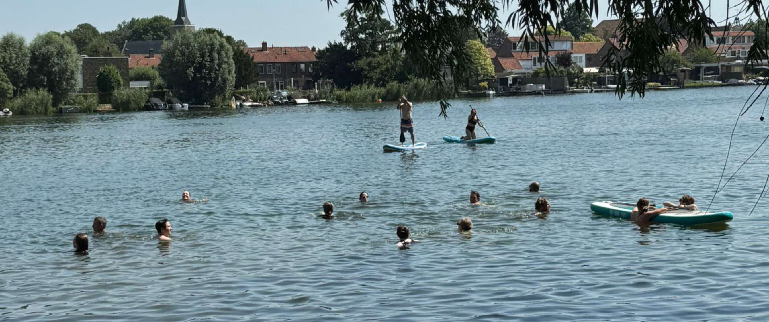 Des nageurs et deux personnes sur paddle évoluent sur une eau calme près d’une rive bâtie.