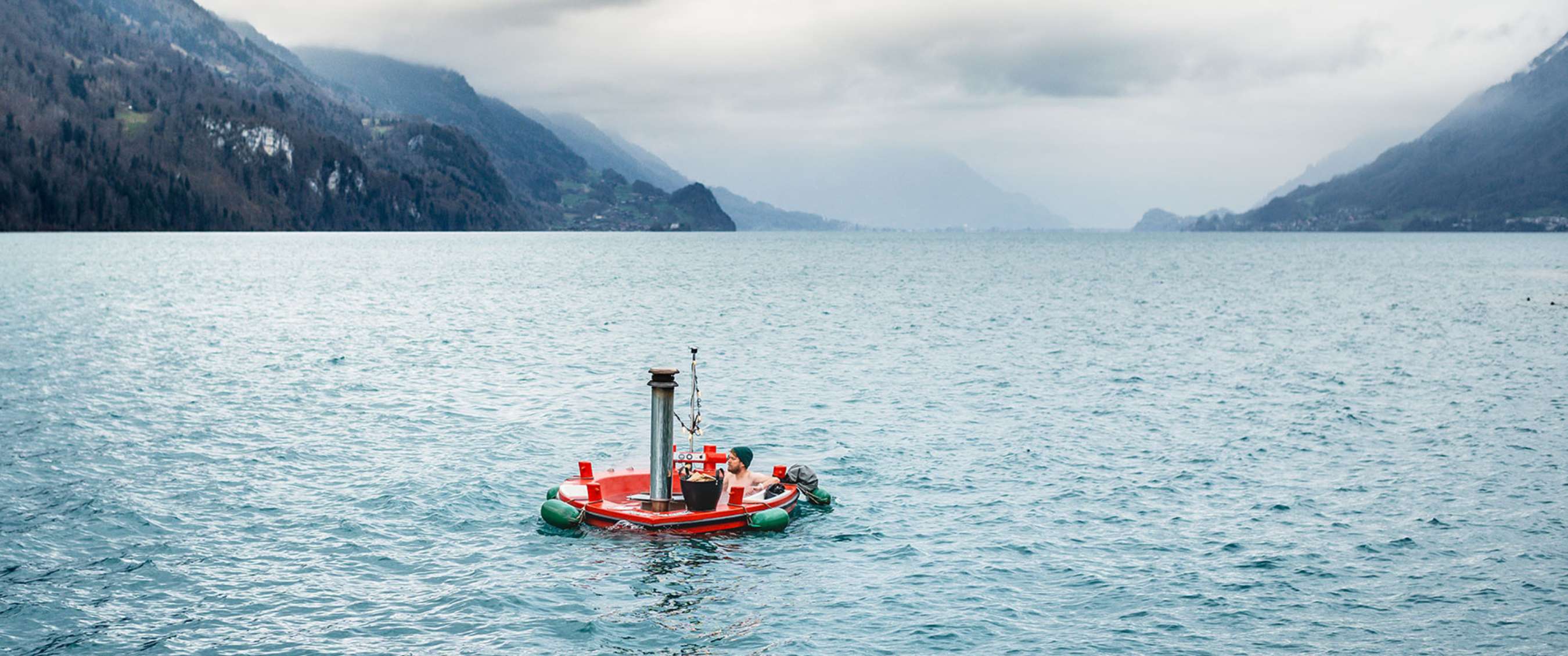 Plateforme rouge flottante avec une personne sur un grand lac entouré de montagnes sous un ciel couvert.