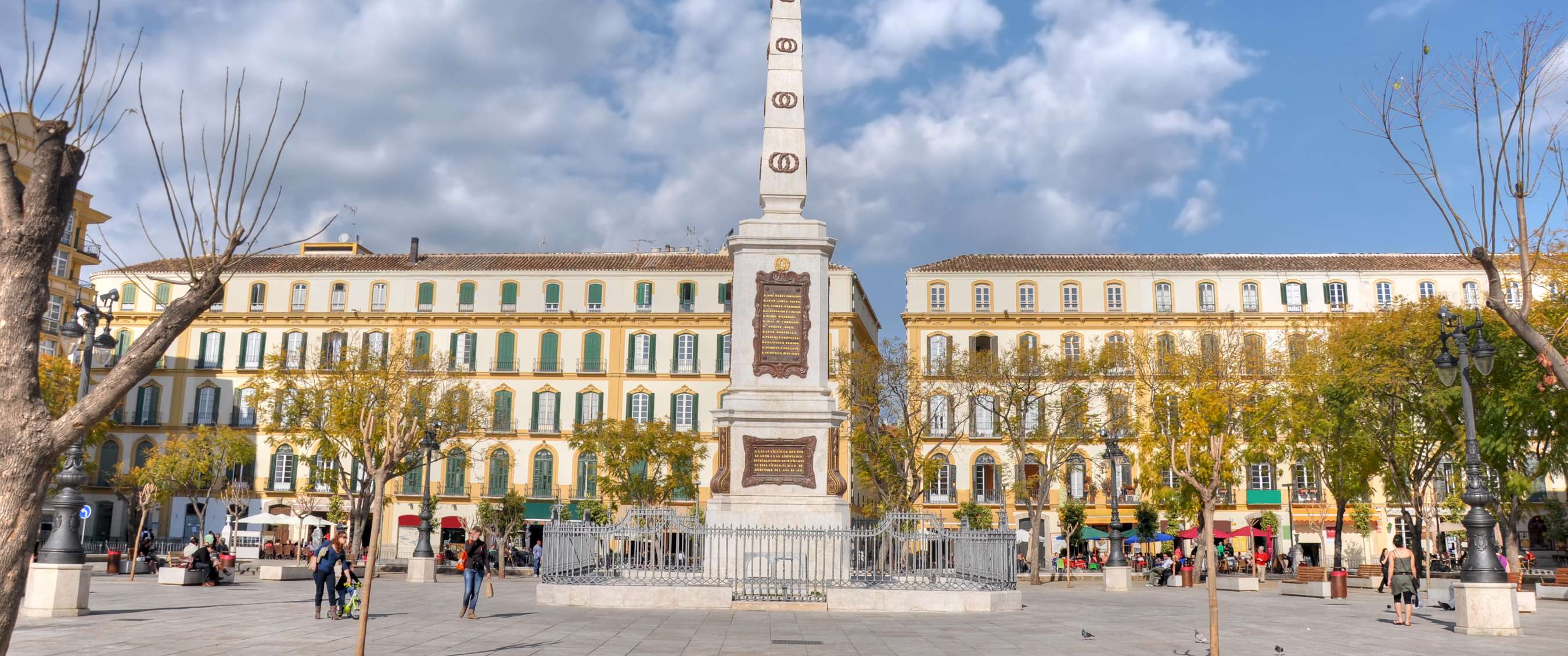 Obélisque blanc sur une vaste place de Málaga, entouré de façades historiques et d’arbres.