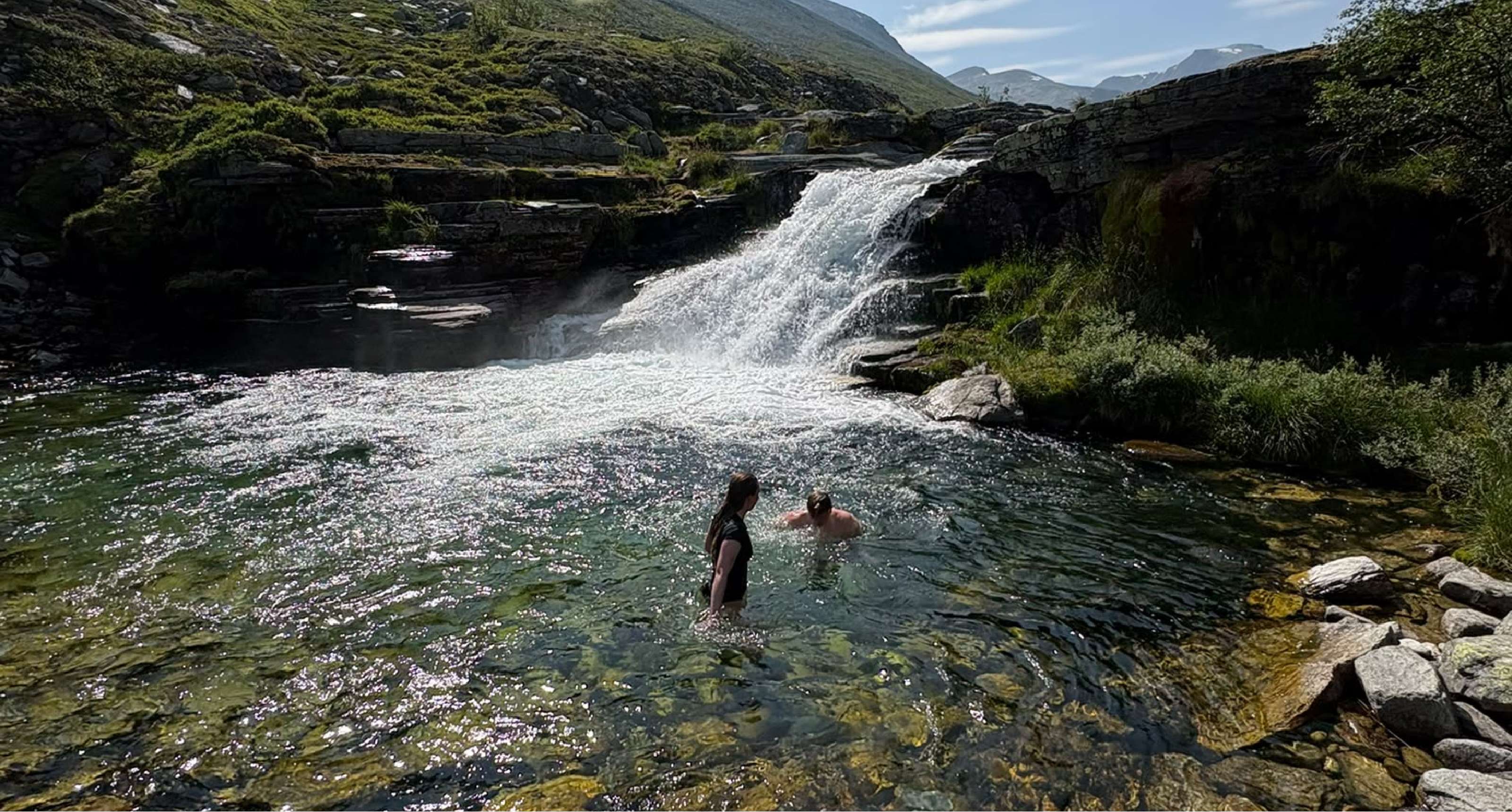 Deux personnes se tiennent dans une eau claire sous une cascade, entourées de rochers et de végétation.