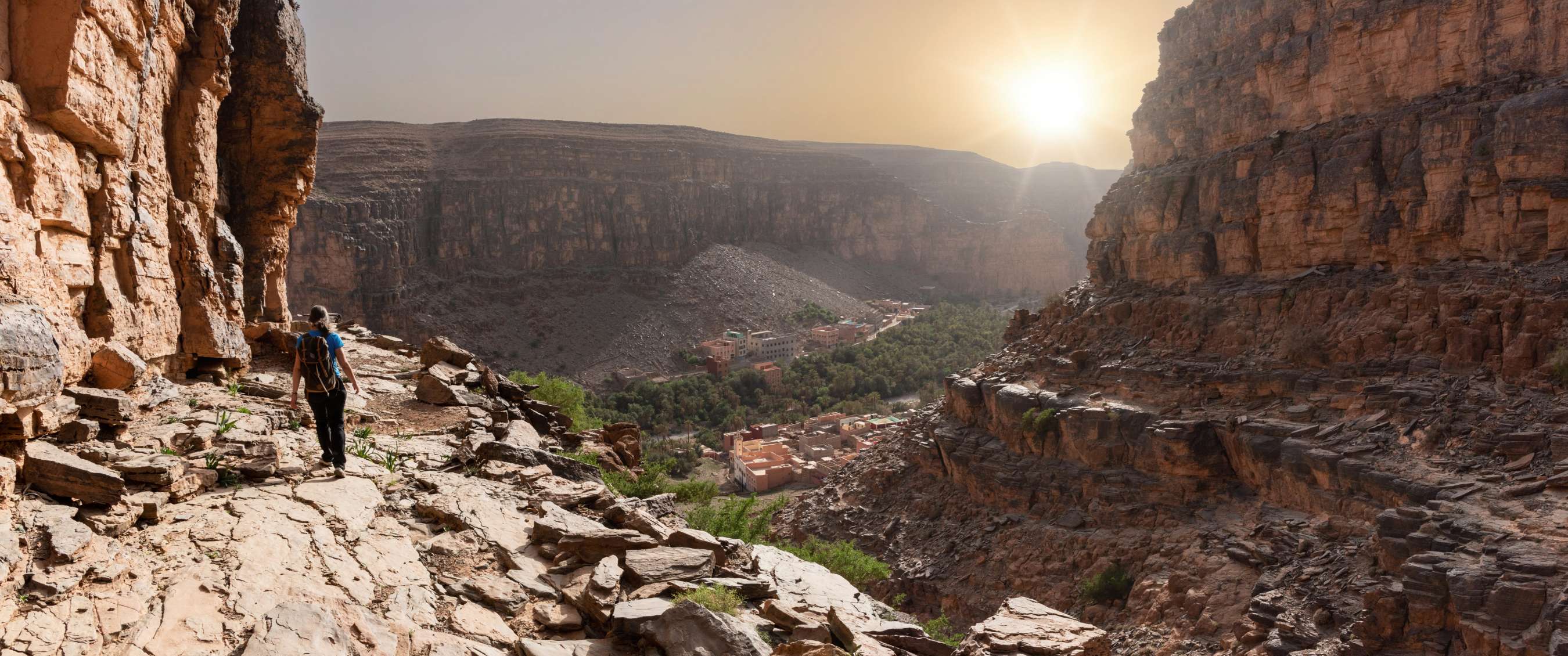 Randonneur avec sac à dos sur un sentier rocheux étroit dans une gorge avec vue sur un village de montagne au soleil bas.