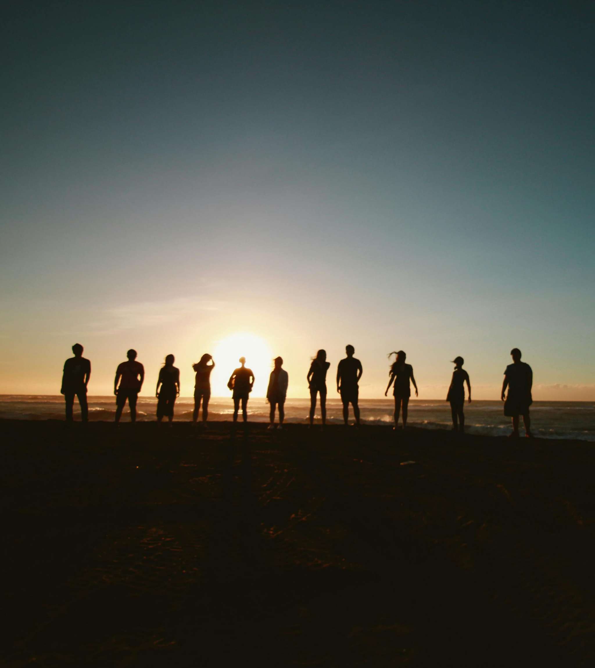 Groupe de personnes en silhouettes sur la plage avec le soleil bas à l’horizon.
