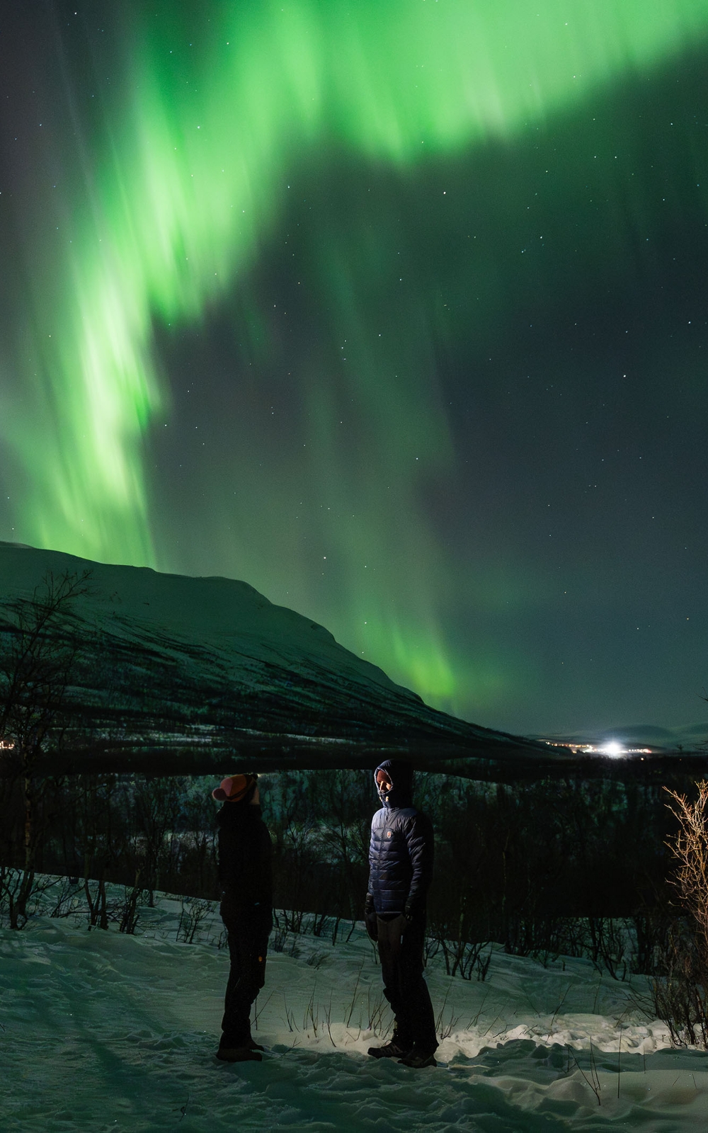 Aurore boréale verte au-dessus de montagnes enneigées avec deux personnes dans l’obscurité.