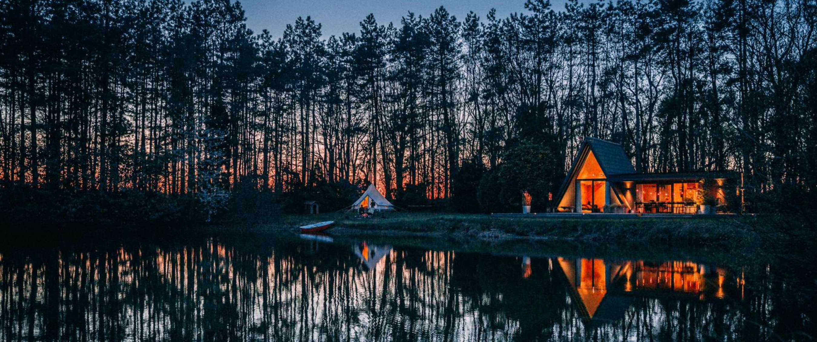 Maison de vacances éclairée au bord de l’eau au crépuscule, entourée d’arbres et reflétée dans le lac.