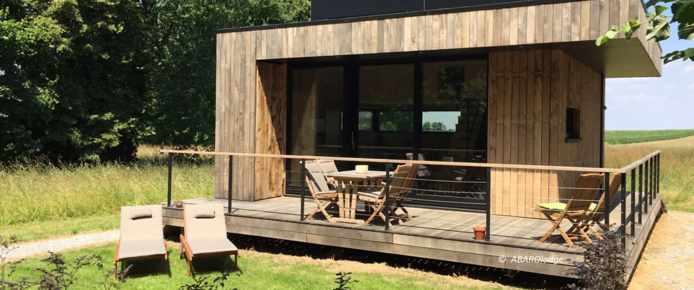 Maison de vacances moderne en bois avec terrasse surélevée, chaises et transats dans un cadre verdoyant.