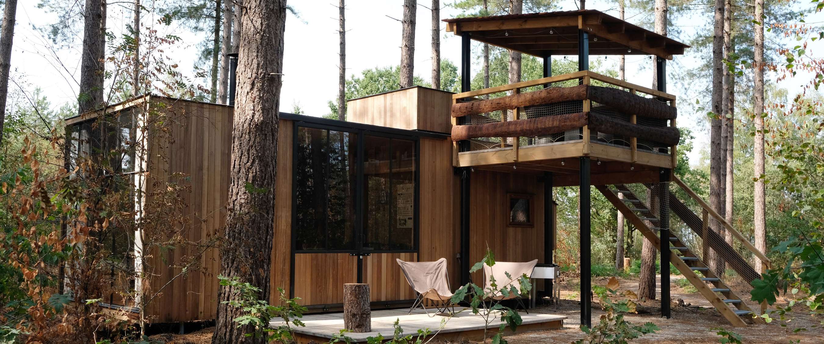 Maison de vacances en bois parmi les arbres avec terrasse surélevée et plateforme accessible par un escalier.
