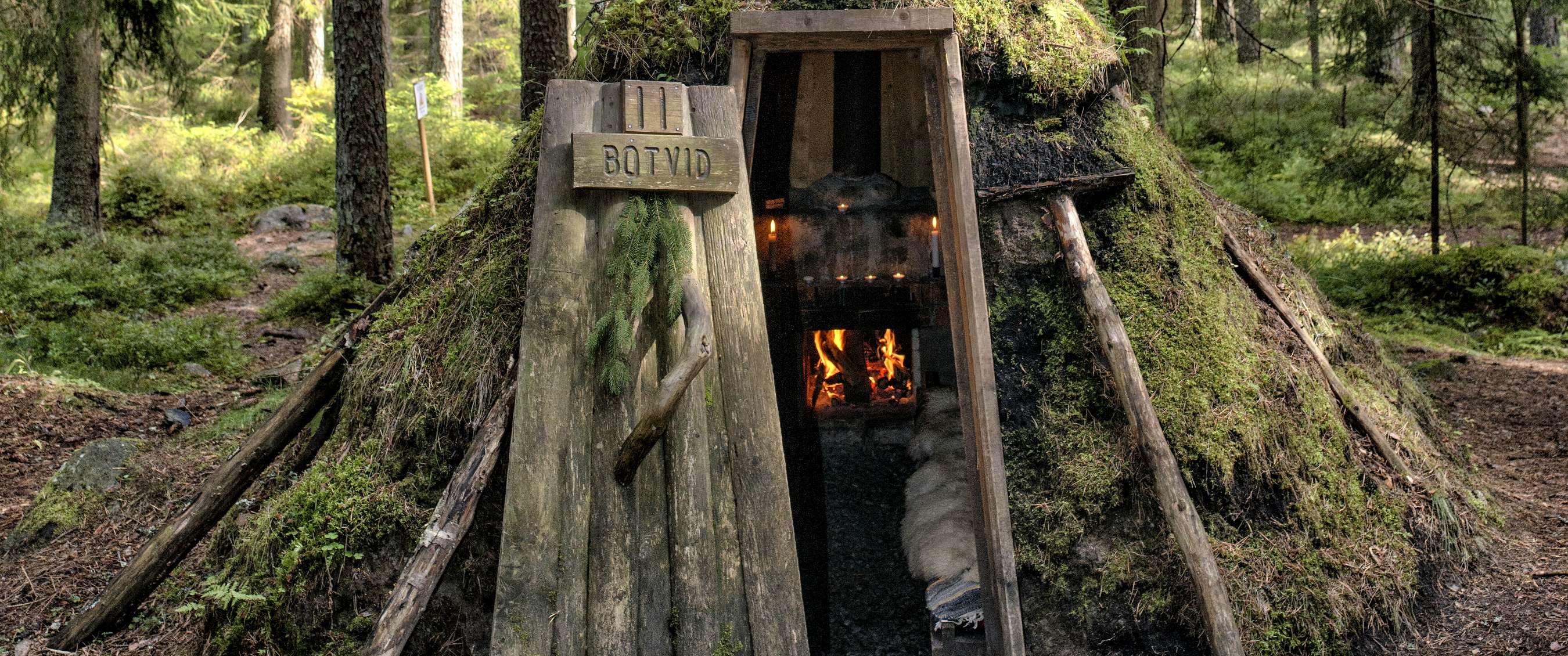 Abri en bois recouvert de mousse dans la forêt, avec porte ouverte, feu à l’intérieur et panneau “BOTVID”.