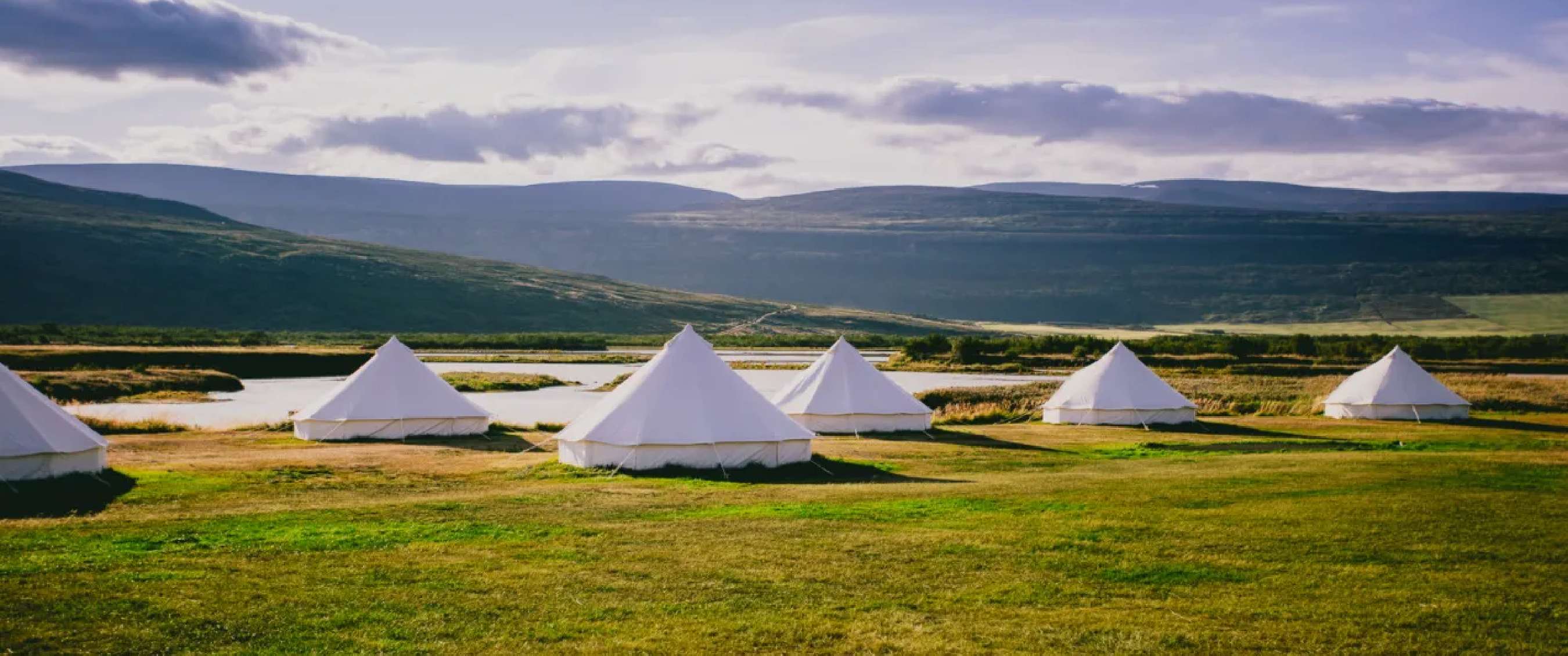 Tentes de glamping blanches installées dans un paysage herbeux ouvert, avec rivière et montagnes en arrière-plan.