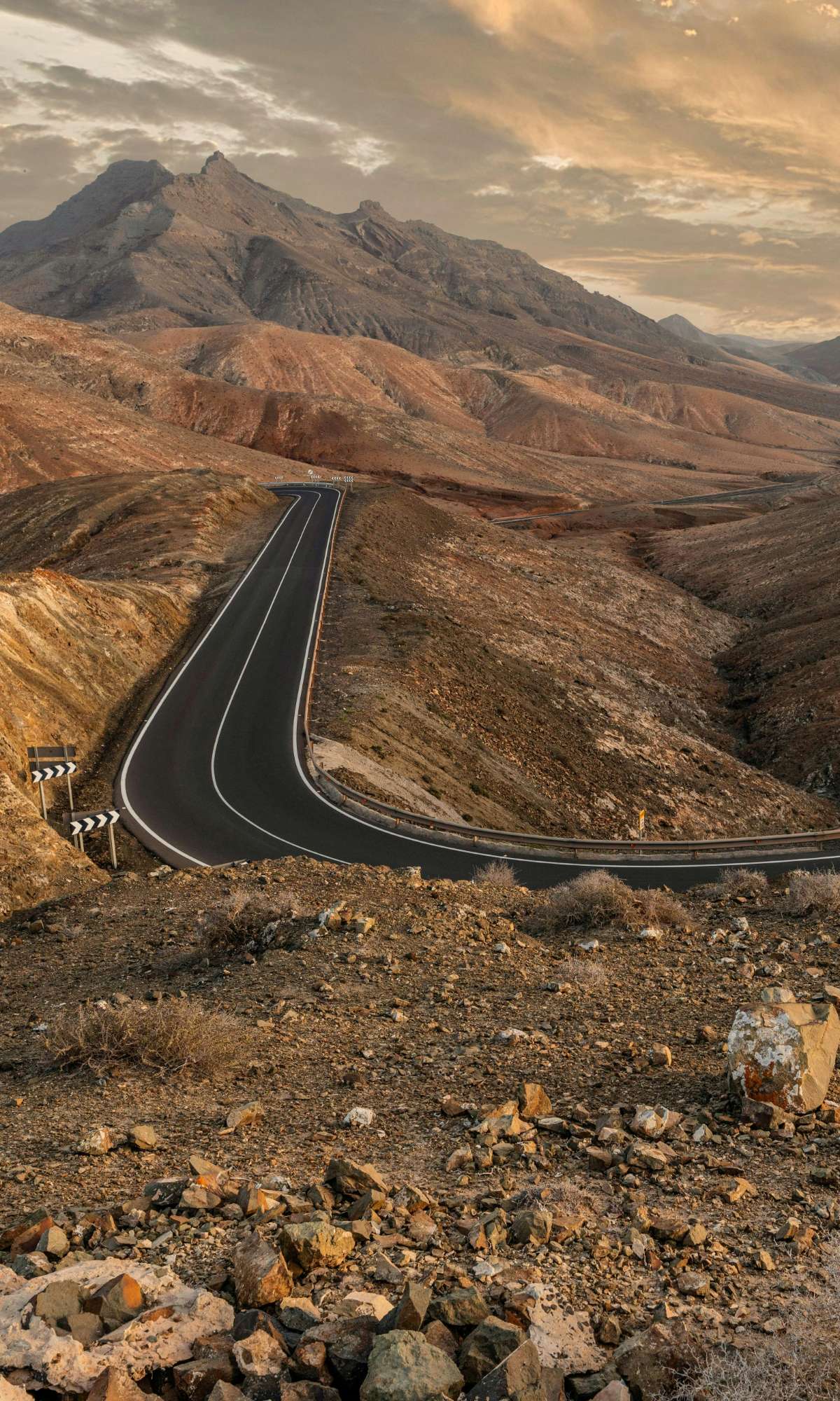 Route asphaltée sinueuse dans un paysage montagneux aride au coucher du soleil.