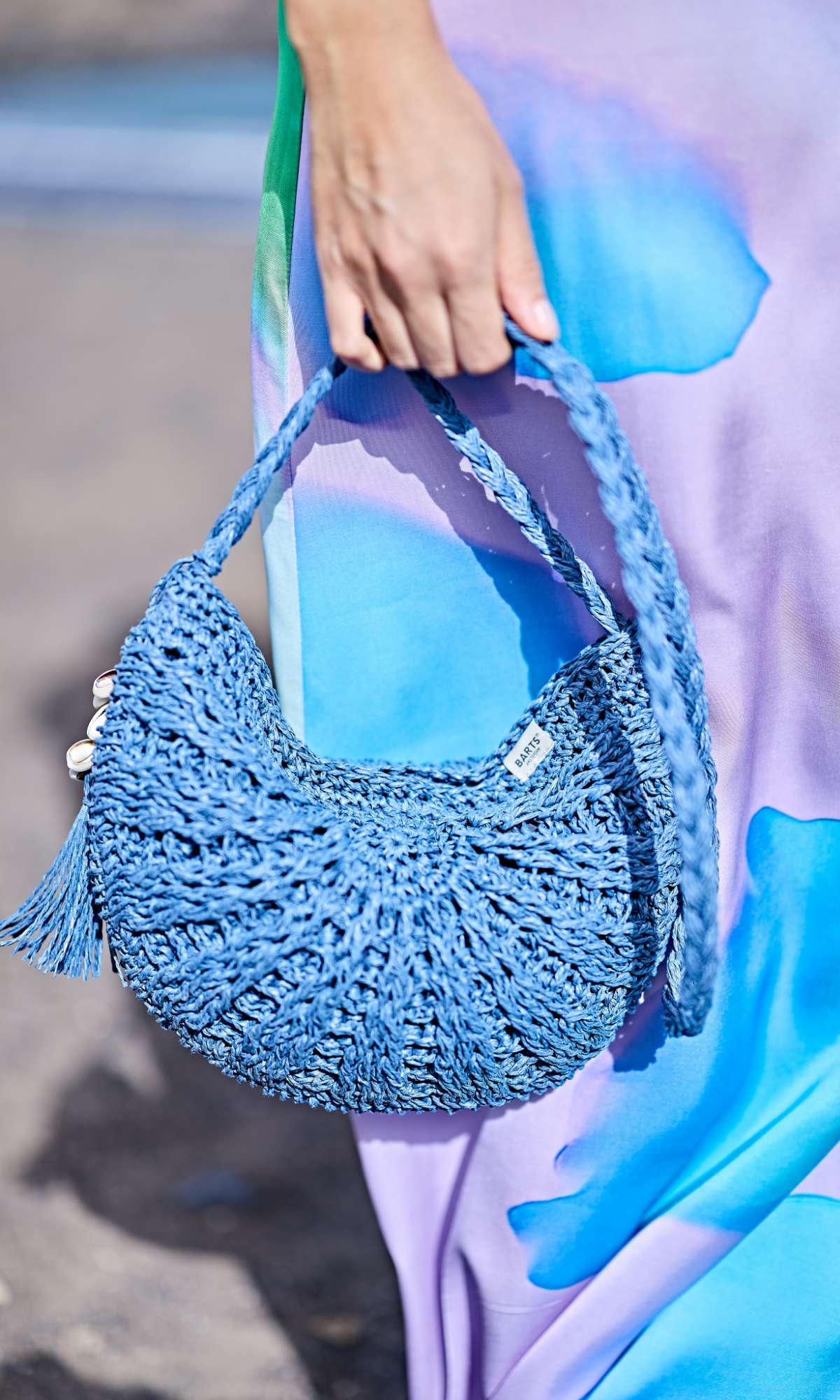 Main tenant un sac à main bleu en crochet devant une robe violette à imprimé bleu.