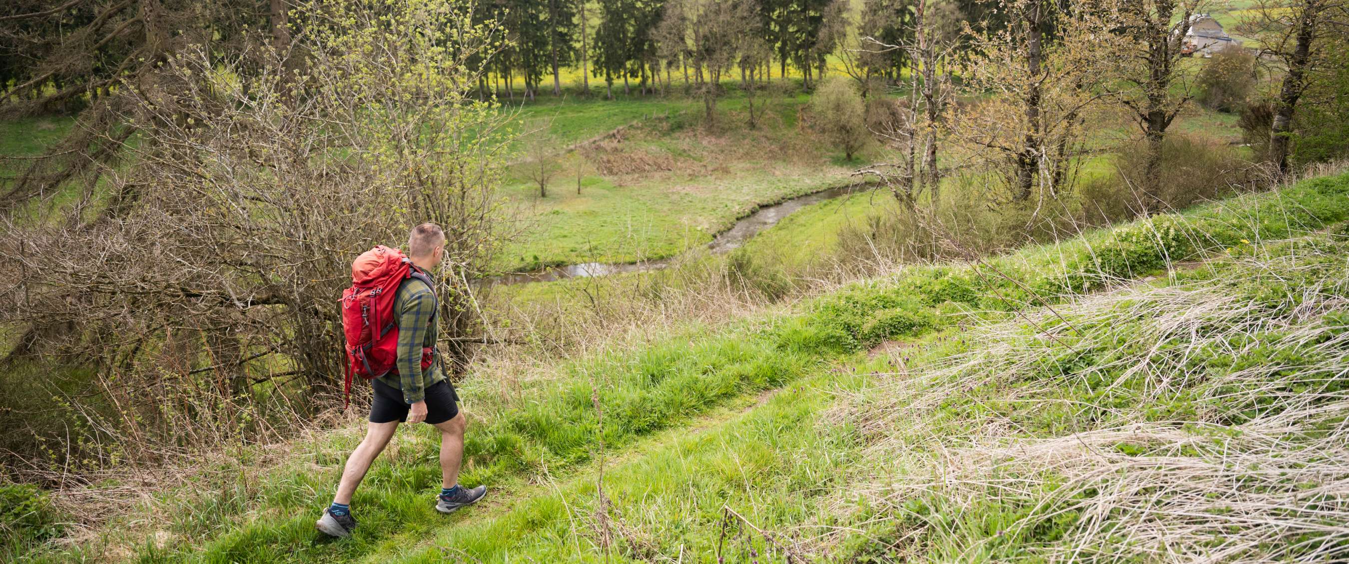 Personne avec sac à dos marchant dans l’herbe près d’un ruisseau vallonné.