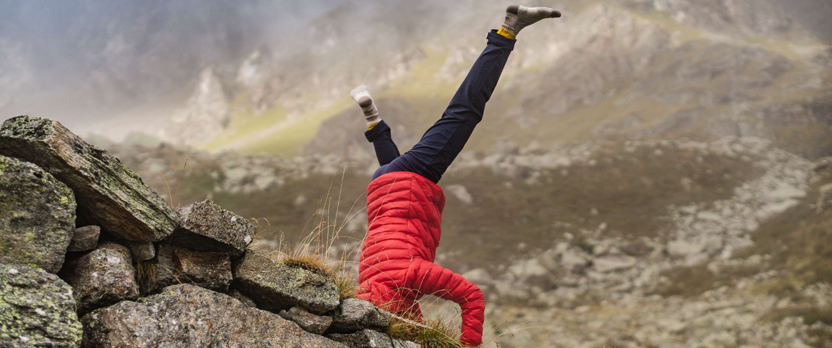 Une personne en veste rouge fait un équilibre sur les mains en milieu alpin.
