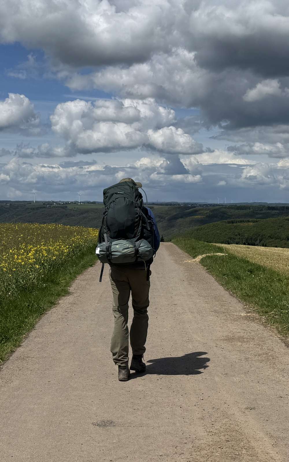 Randonneur avec grand sac sur un large chemin rural sous un ciel nuageux