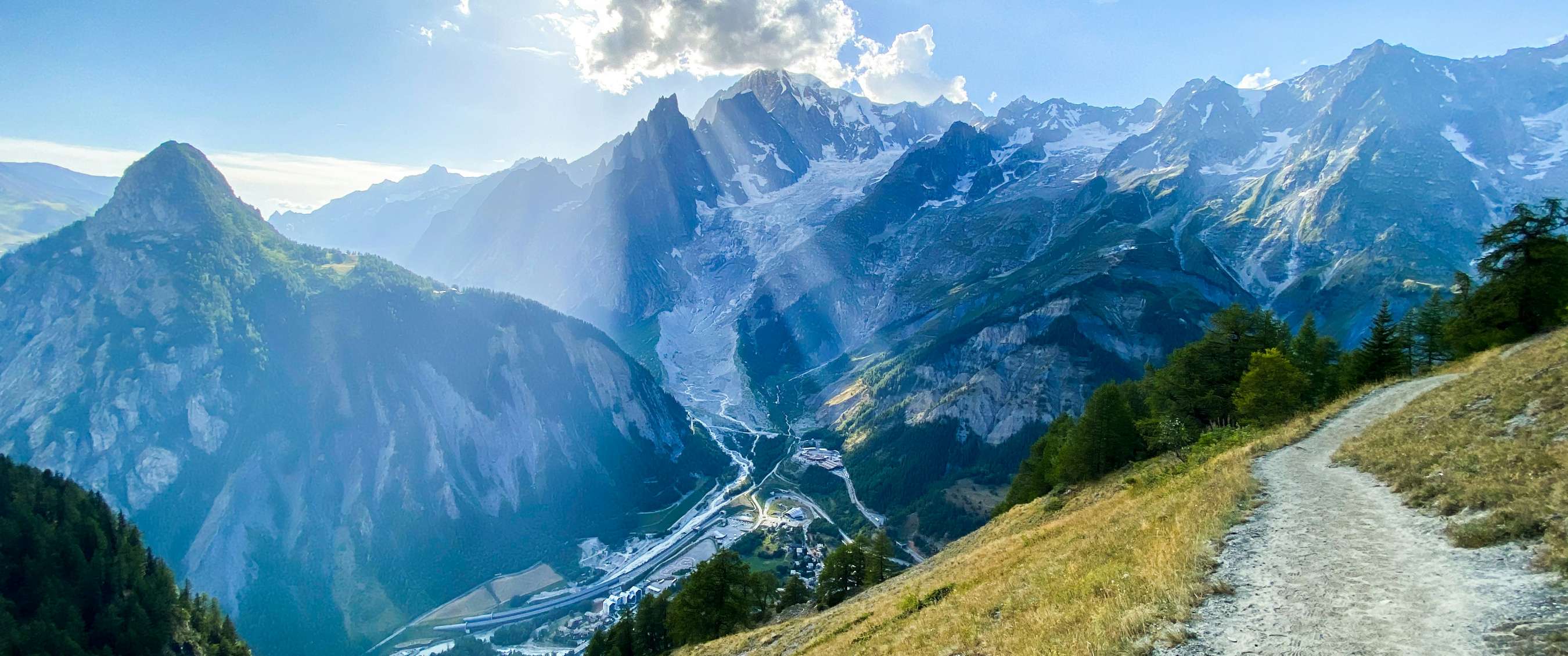 Panorama de montagne avec sentier étroit, glacier et soleil au-dessus des sommets.