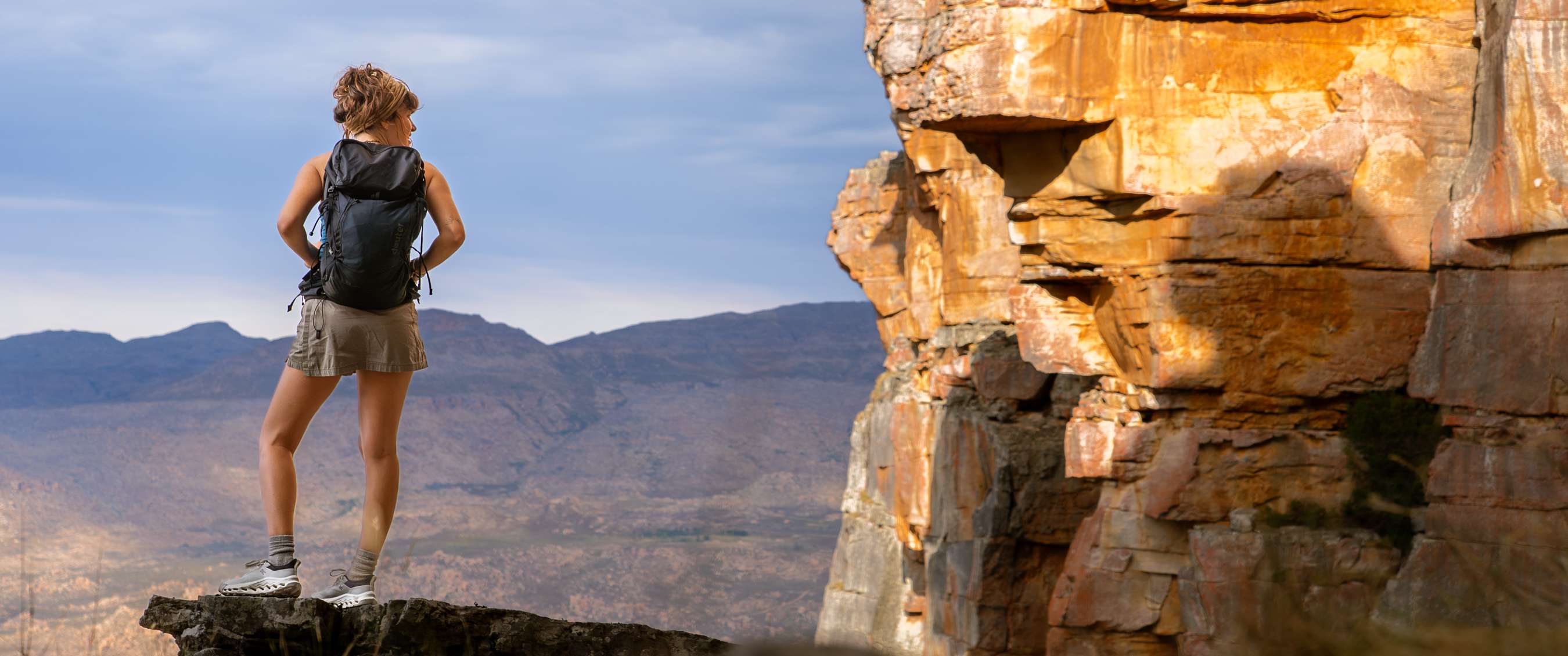 Randonneuse sur un rocher en surplomb avec panorama de montagnes et lumière sur la paroi voisine.