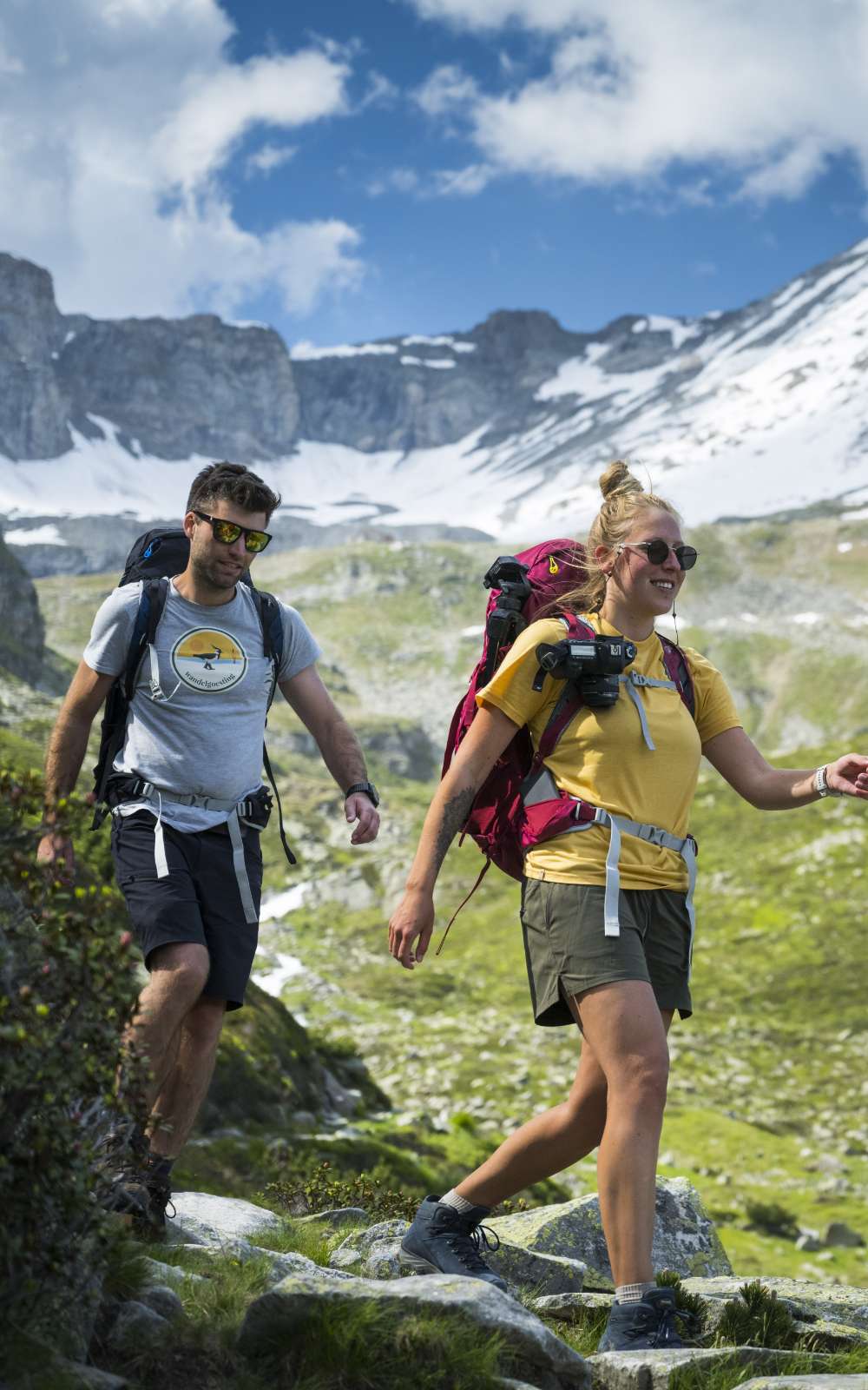 Deux randonneurs avec sacs descendent un sentier rocheux avec neige en arrière-plan.
