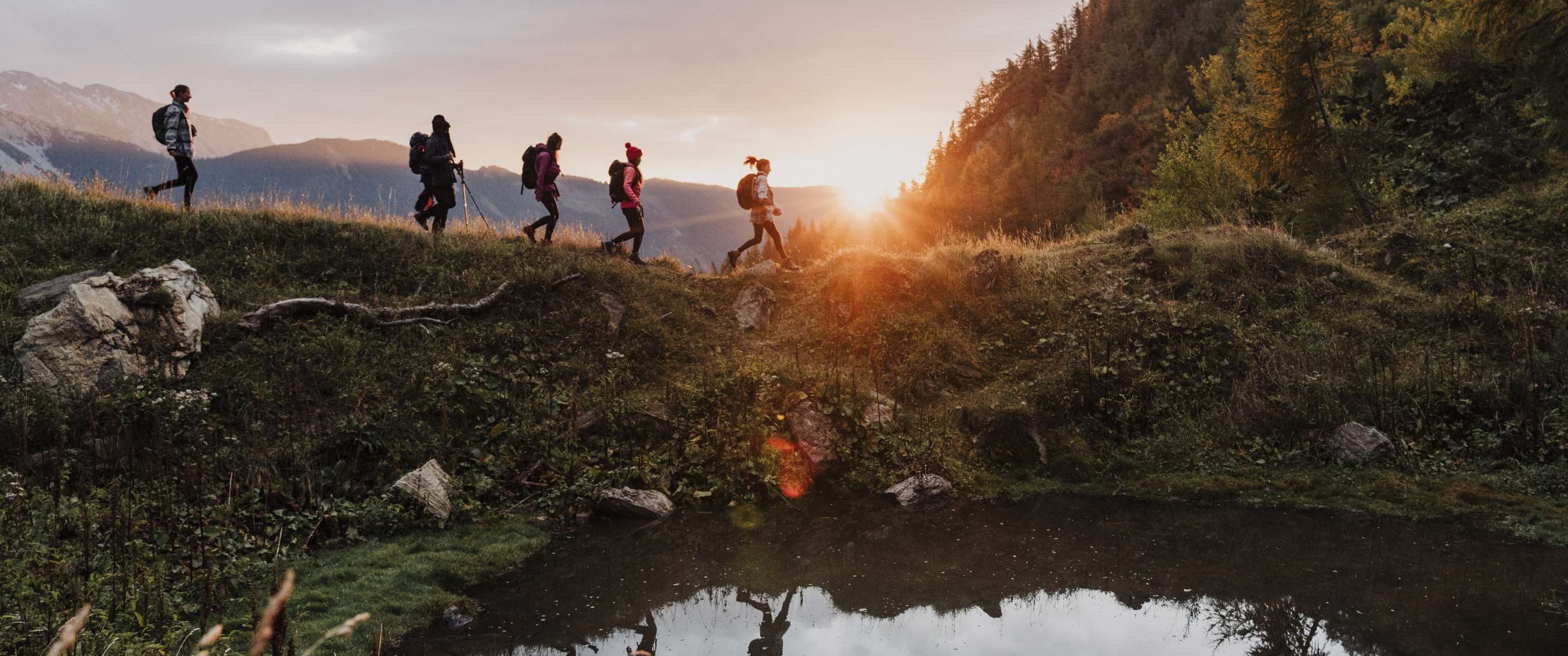Groupe de randonneurs avec sacs marchant près de l’eau au coucher du soleil.