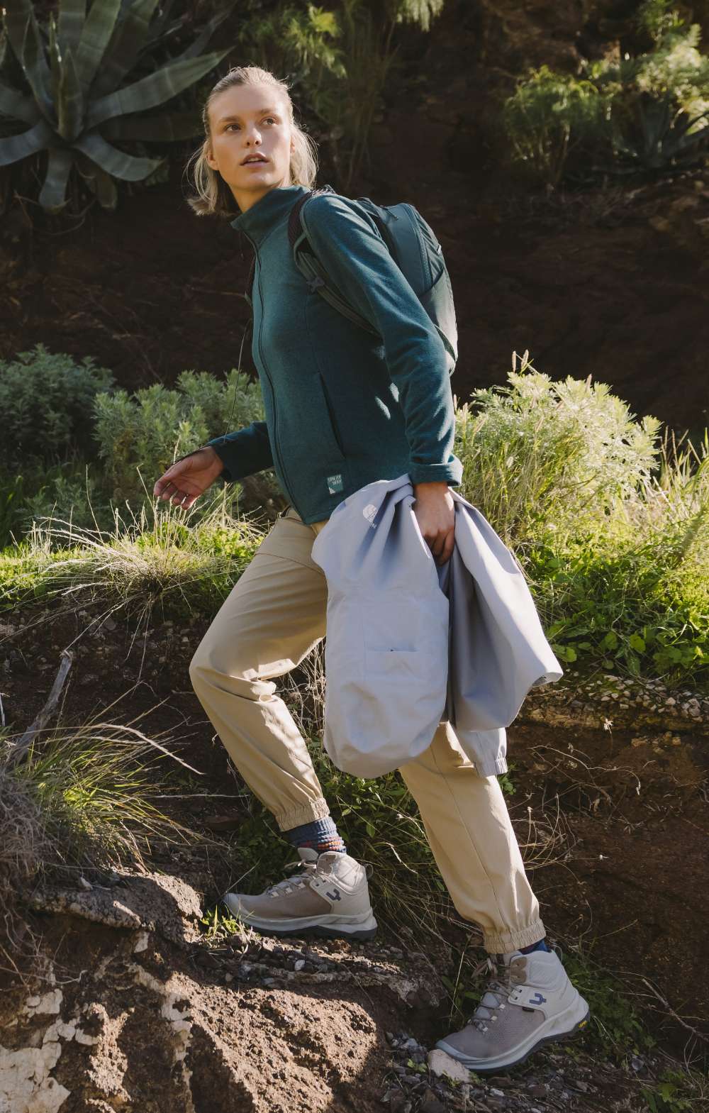 Femme en pantalon de randonnée beige et veste polaire bleue marchant sur un sentier rocheux avec une veste à la main