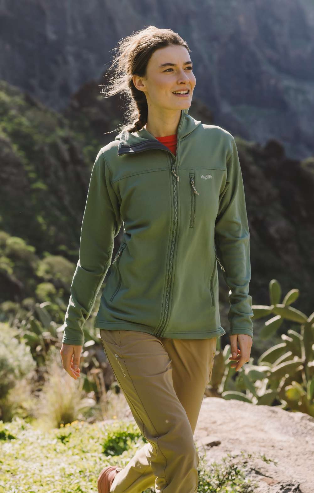 Femme en veste verte et pantalon de randonnée beige marchant dans un paysage montagneux ensoleillé
