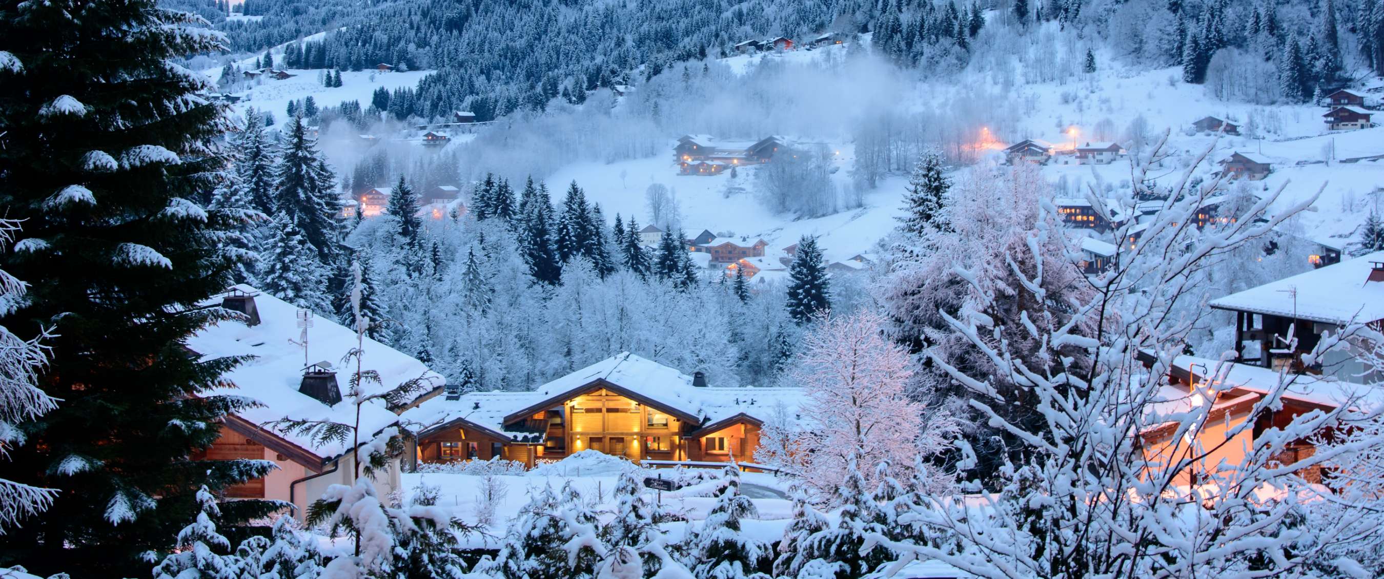 Chalets enneigés entre sapins avec éclairage du soir.