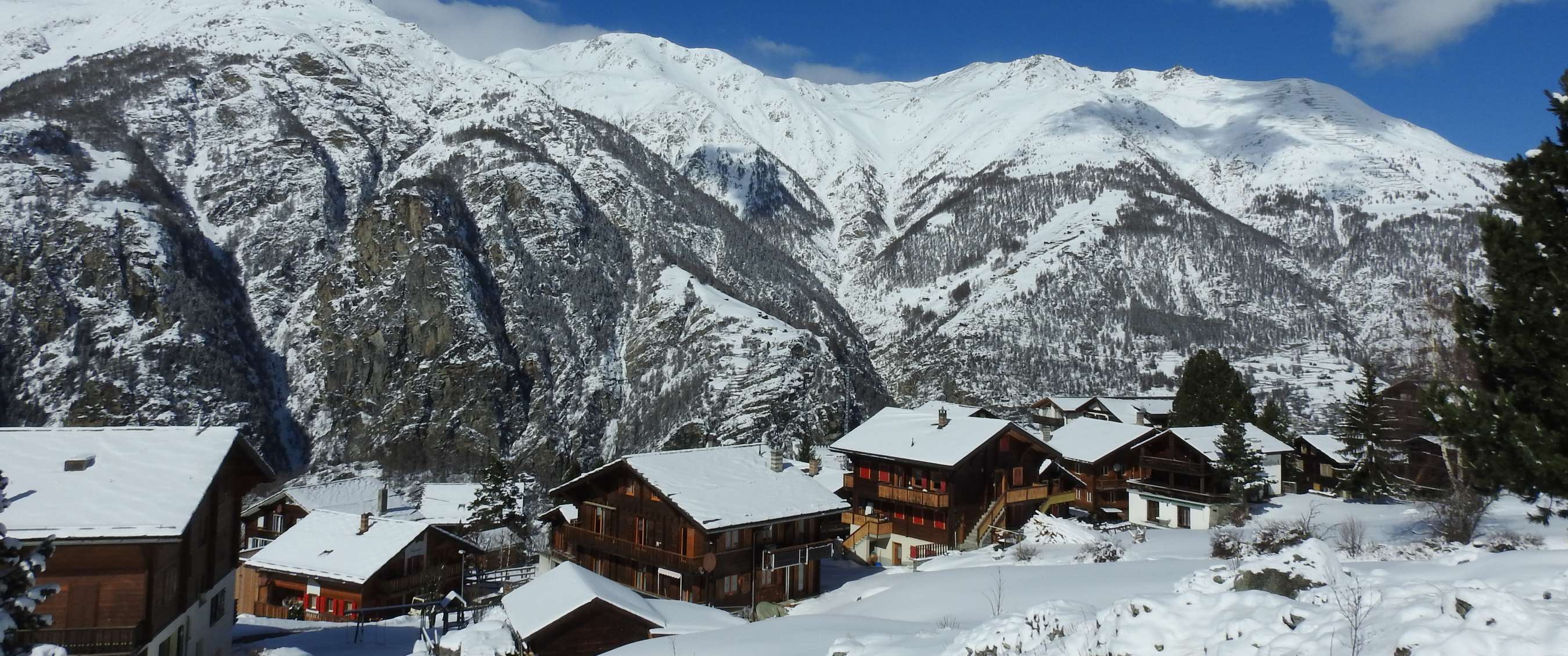 Village alpin avec chalets en bois devant parois montagneuses enneigées.