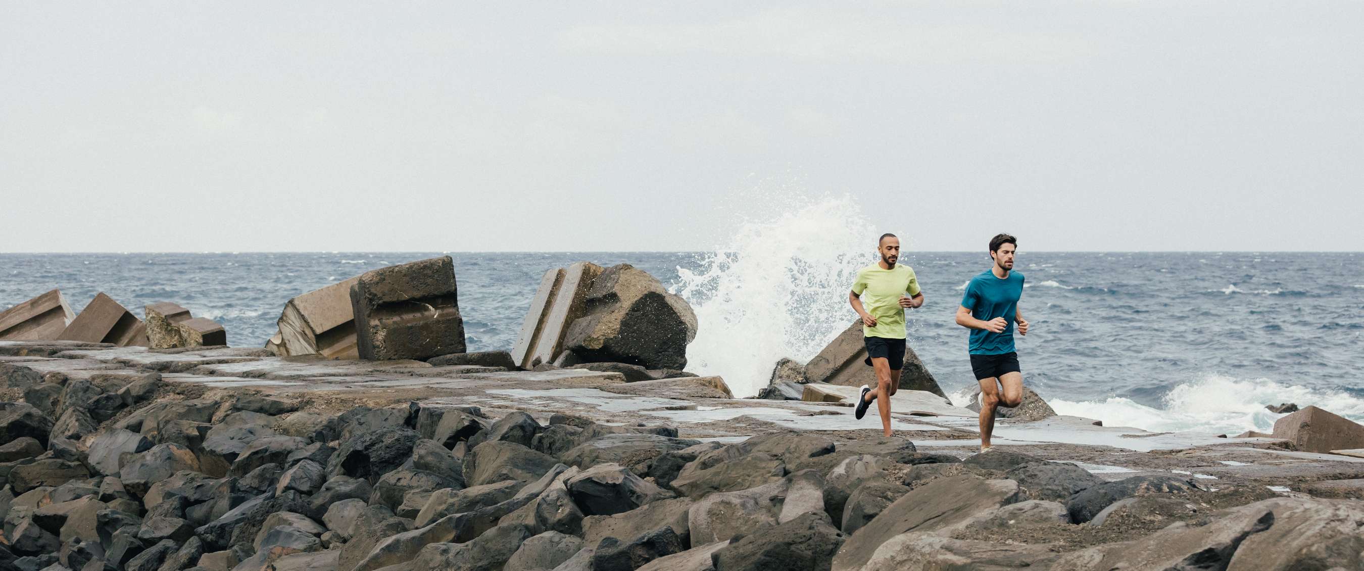 Deux coureurs longent des rochers près de la mer avec des vagues qui éclaboussent.