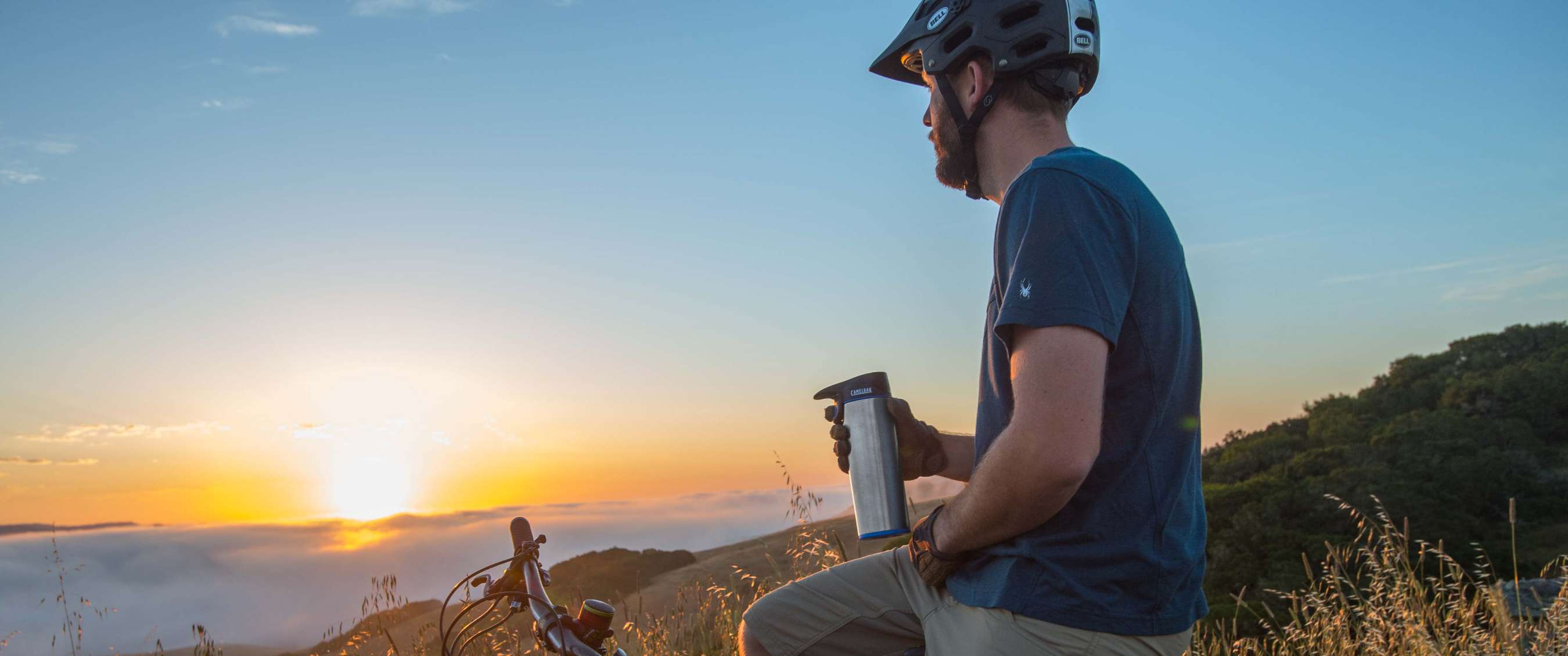 Un cycliste casqué est assis près d’un VTT et tient une gourde métallique au coucher du soleil.
